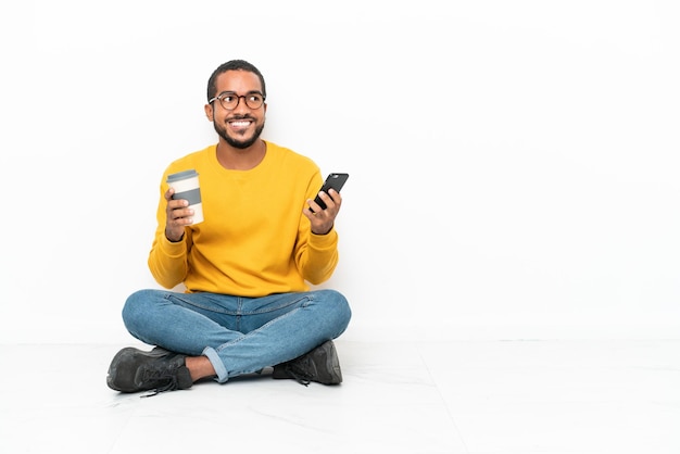 Young Ecuadorian man sitting on the floor isolated on white wall holding coffee to take away and a mobile while thinking something