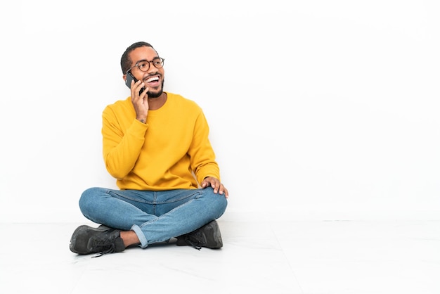 Young Ecuadorian man sitting on the floor isolated on white wall keeping a conversation with the mobile phone