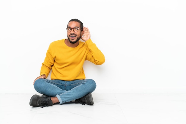 Young Ecuadorian man sitting on the floor isolated on white wall listening to something by putting hand on the ear