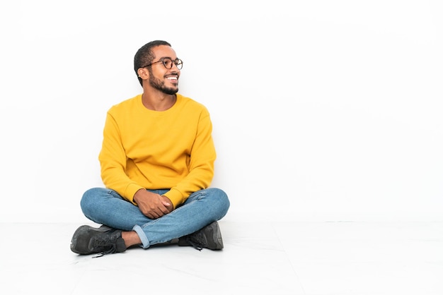 Young Ecuadorian man sitting on the floor isolated on white wall looking side