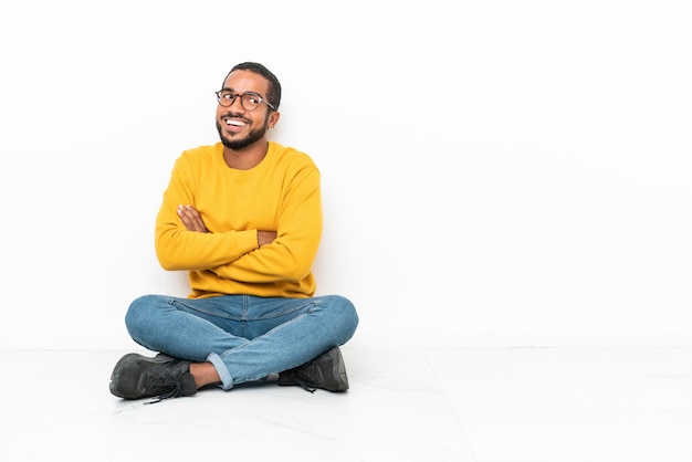 Young Ecuadorian man sitting on the floor isolated on white wall with arms crossed and happy