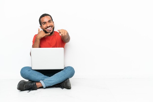 Young Ecuadorian man with a laptop sitting on the floor isolated on white background making phone gesture and pointing front