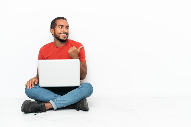 Young Ecuadorian man with a laptop sitting on the floor isolated on white background pointing to the side to present a product