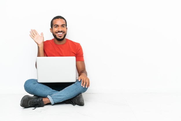 Giovane uomo ecuadoriano con un computer portatile seduto sul pavimento isolato su sfondo bianco salutando con la mano con un'espressione felice