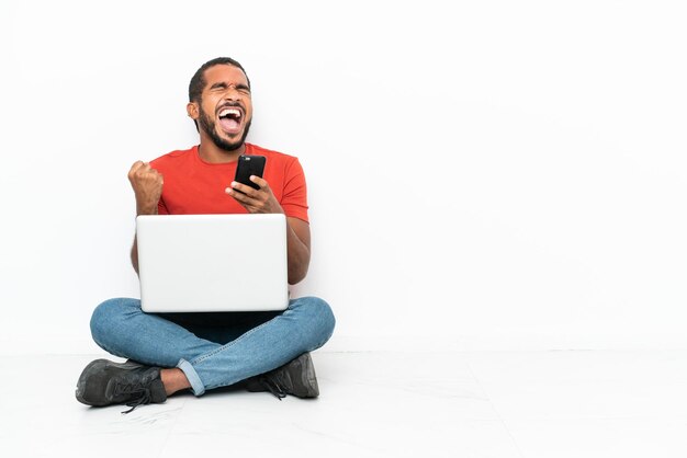 Young Ecuadorian man with a laptop sitting on the floor isolated on white background with phone in victory position