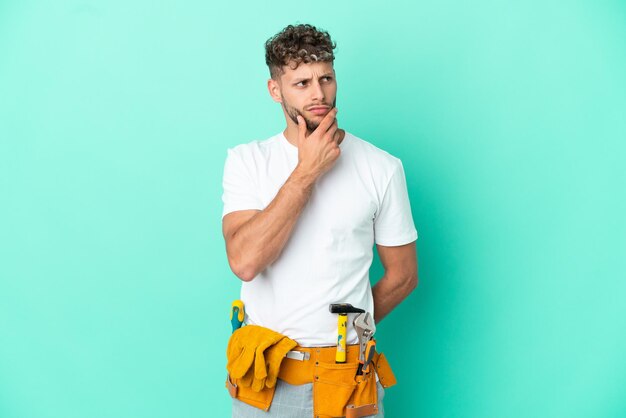 Young electrician blonde man isolated on green background having doubts and with confuse face expression