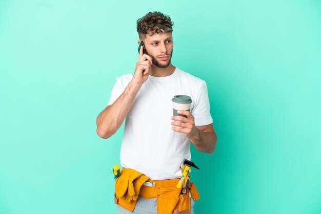 Young electrician blonde man isolated on green background holding coffee to take away and a mobile