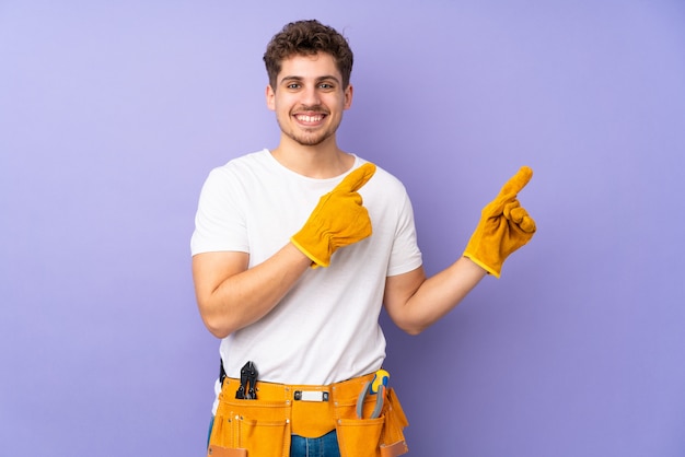 Young electrician man over isolated on purple wall pointing finger to the side