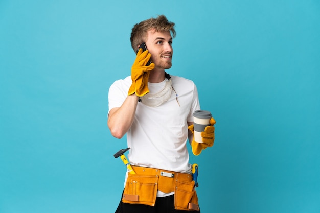 Young electrician man over isolated wall holding coffee to take away and a mobile