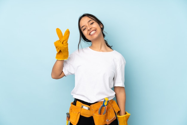 Young electrician woman on blue smiling and showing victory sign