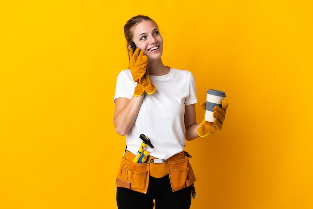 Young electrician woman isolated on yellow holding coffee to take away and a mobile