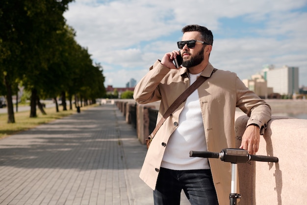 Young elegant agent with electric scooter talking on mobile phone in urban environment while standing between riverside and park