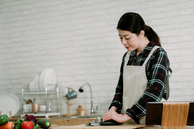 Young elegant asian korean woman in modern kitchen following\
recipe online website on digital tablet. smiling wife in apron\
chopping cucumber with sharp knife on wooden cutting board in\
cooking place