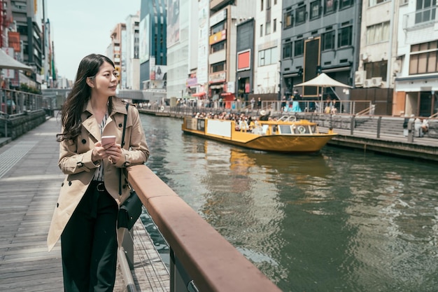 오사카 시의 수로 근처에 서 있는 가방을 들고 스마트폰을 사용하는 젊고 우아한 아시아 여성