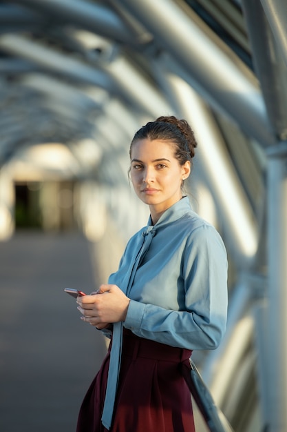 Young elegant brunette female delegate looking at you while scrolling in smartphone in front of camera inside contemporary business center