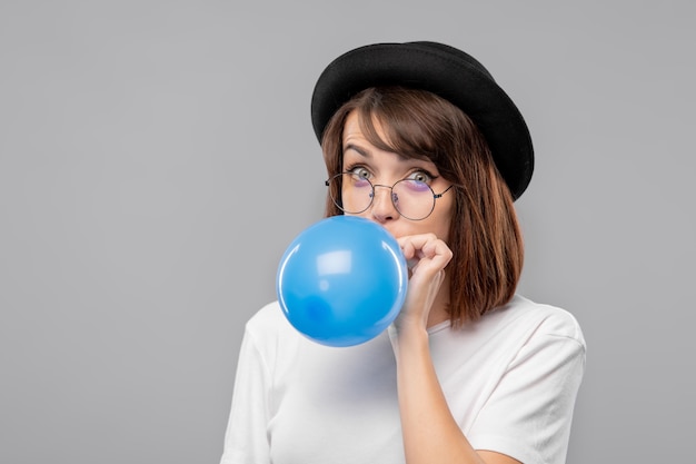 Young elegant brunette female in white t-shirt and black hat blowing blue balloon in isolation