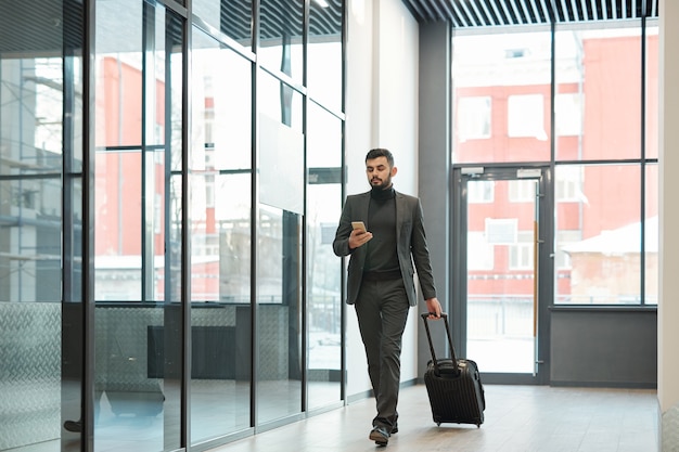 Молодой элегантный бизнесмен прокручивает смартфон во время движения по аэропорту и тянет чемодан с багажом