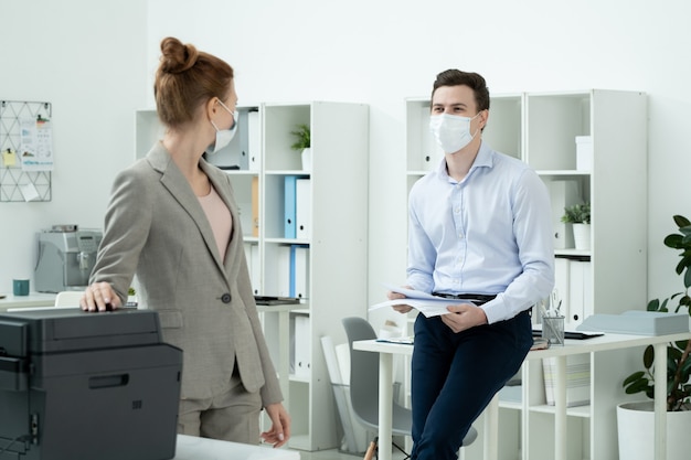 Young elegant businessman with papers standing by desk in front of his female colleague using xerox machine during conversation in office