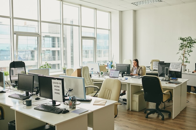 Photo young elegant businesswoman sitting by desk in the corner of modern office and looking at computer screen during pandemic period