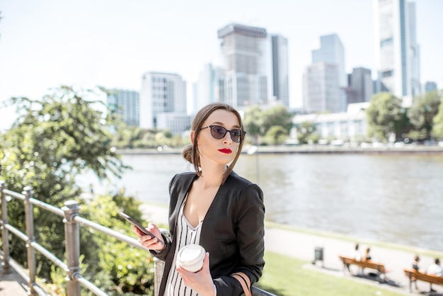 Young and elegant businesswoman with phone and coffee standing on the quay with skyscrapers on the background in Frankfurt city