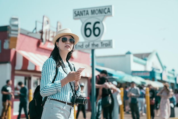 young elegant female tourist with sunglasses and hat holding mobile phone standing in amusement park by Santa Monica 66 sign California USA. asian japanese woman carry professional camera travel