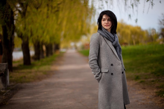 Young elegant girl in black glasses, a gray coat and a gray scarf on a walk in the park by the lake