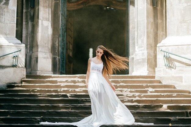 Young  elegant girl with flying hair in long white dress posing on the stairs of ancient palace