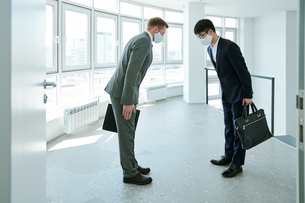 Young elegant intercultural businessmen with briefcases greeting each other by bow at meeting inside large contemporary office