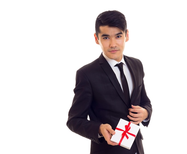 Young elegant man with black hair in white shirt and black tuxedo with black tie holding a present