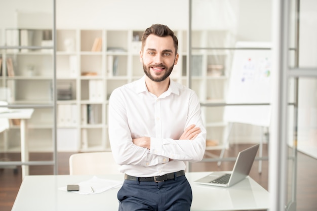 Young elegant office manager or broker crossing his arms on chest while standing by workplace