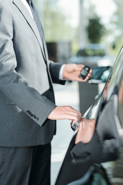 Young elegant seller of new cars in suit using remote control alarm system while standing by one of automobiles and going to open the door