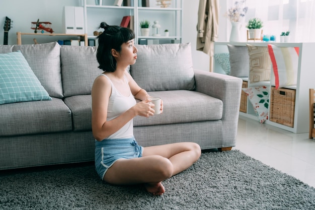 窓から日の出の太陽の光で自宅でコーヒーを飲むコンフォートショーツの若いエレガントな女性。側面図の肖像画家のリビングルームのカーペットの上に座っている美しい女性は、屋内で日光をお楽しみください