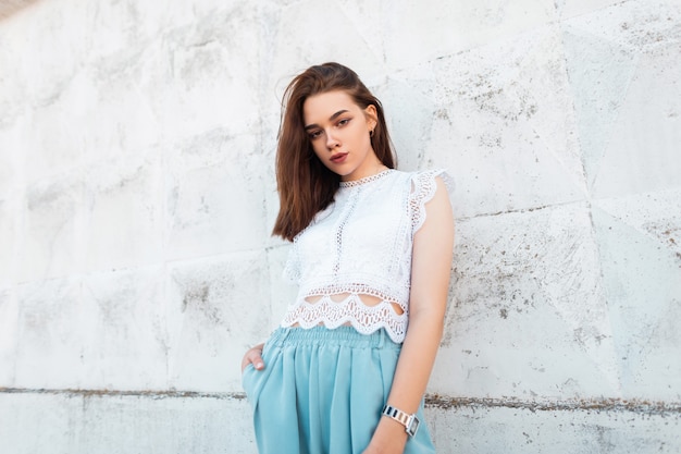 Young elegant woman model in a fashionable lace top in stylish blue pants is standing in the city near a white vintage wall. Attractive fashion model girl in the street. Summer style.