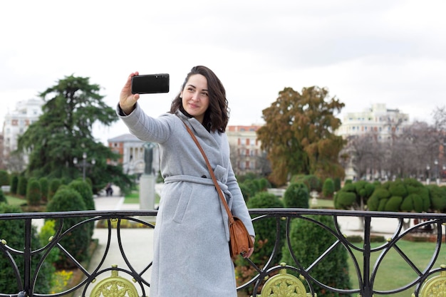 Молодая элегантная женщина осматривает достопримечательности. Она фотографирует себя на мобильный телефон в городском саду.