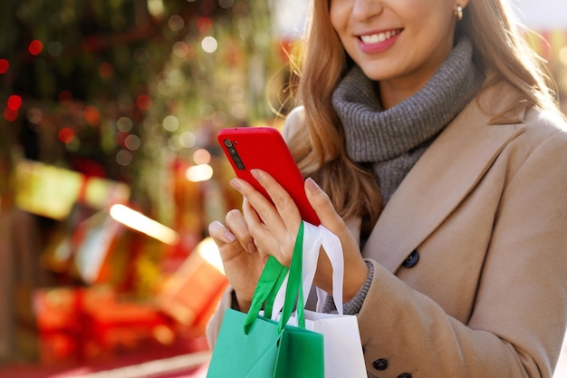 Young elegant woman using smartphone for shopping online and carrying bags on Christmas time. Cropped Christmas Eve picture with focus on the hand and phone.