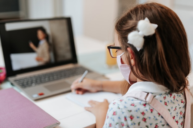 写真 オンライン教育クラスを見ている顔保護マスクを持つ若い小学生の女の子。コロナウイルスまたはcovid-19封鎖教育の概念。