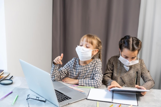 얼굴 보호 마스크를 쓴 어린 초등학교 여학생들이 온라인 교육 수업을 보고 있습니다. 코로나바이러스 또는 코비드-19 잠금 교육 개념.
