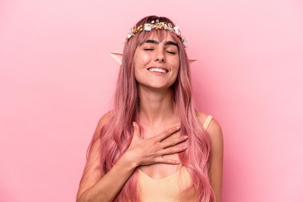 La giovane donna elfo con i capelli rosa isolati su sfondo rosa ride ad alta voce tenendo la mano sul petto.