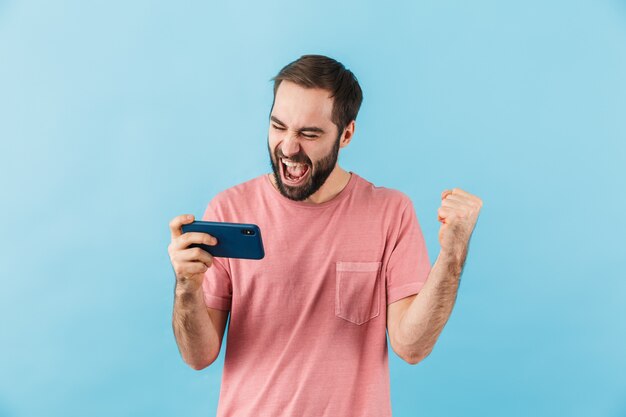 young emotional excited screaming man isolated over blue wall play games by mobile phone make winner gesture.