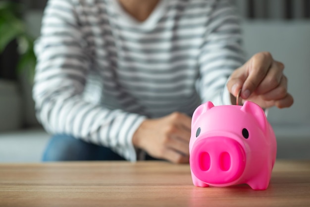 젊은 직원이 동전을 분홍색 돼지 저금통에 넣고 책상에 수입과 지출을 기록하고 있습니다. 그는 미래에 투자하기 위해 친구들의 돈을 저축했습니다