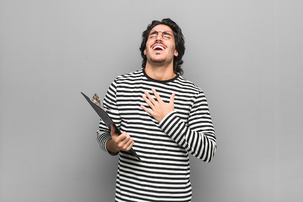 Young employee man holding an inventory laughs out loudly keeping hand on chest.