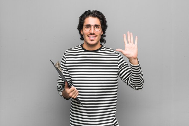 Young employee man holding an inventory smiling cheerful showing number five with fingers