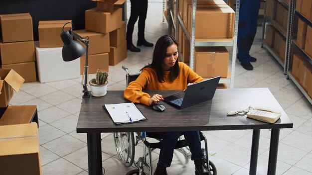 Young employee with impairment working in warehouse, doing desk work for stock logistics and inventory on laptop. Woman wheelchair user doing financial planning in disability friendly space.