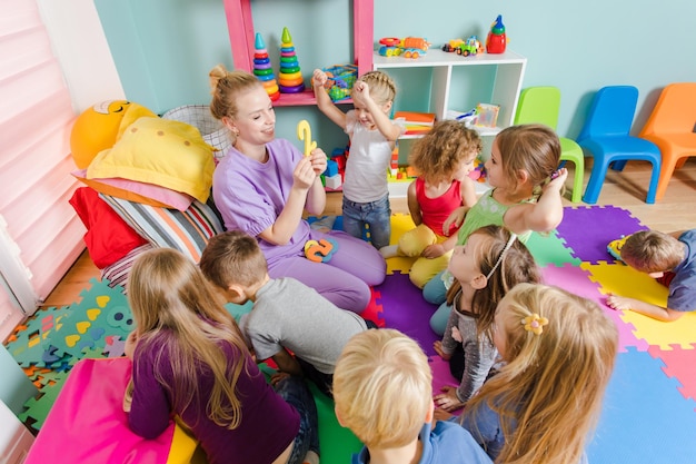 Giovane insegnante impegnato seduto con un gruppo di bambini in età prescolare su un pavimento durante la lezione il processo di apprendimento può essere divertente stanza con tappetini colorati su un pavimento e mobili multicolori