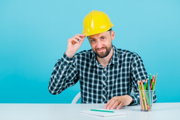 Young engineer man is looking at camera by holding his helmet on blue background