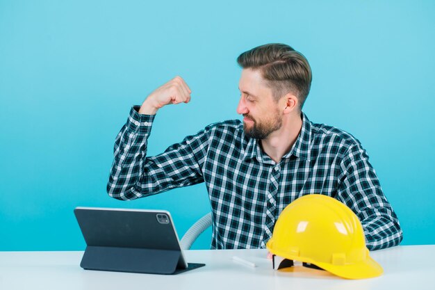 Young engineer man is raising up his muscle by sitting in front of tablet on blue background