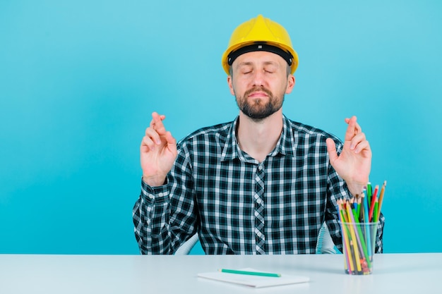 Young engineer man is wishing by crossing fingers on blue background