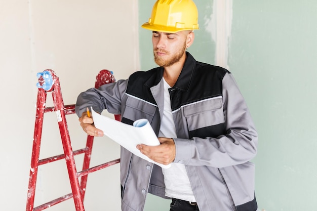 Young engineer in work clothes and yellow hardhat thoughtfully looking on plan of new apartments leaning on red ladder in repairing flat