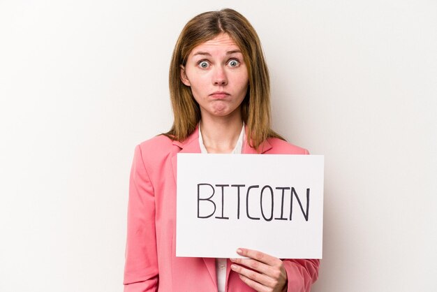 Young English business woman holding a bitcoin placard isolated on white background shrugs shoulders and open eyes confused