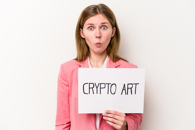 Young English business woman holding a crypto money placard isolated on white background shrugs shoulders and open eyes confused